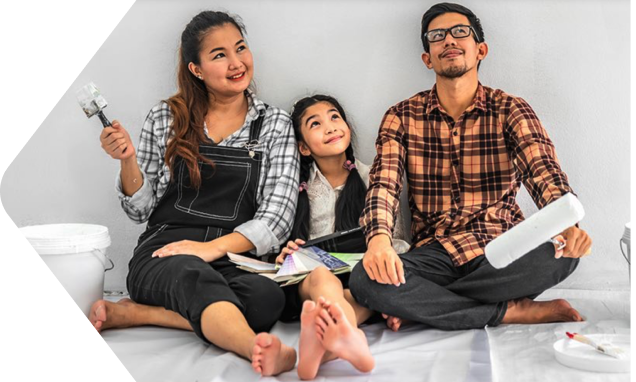 Two young parents hold a paint brush and roller and look up happily at the room they’re painting. A young child sits between them.